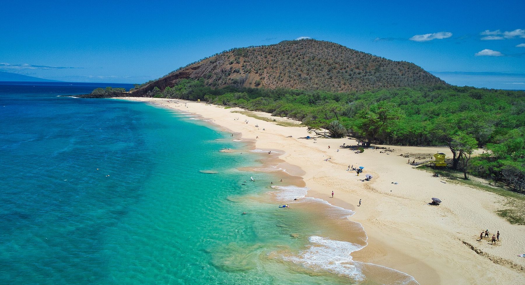 A wide sandy beach with gentle surf and turquoise water curves along a green coastline beneath a rocky hill in Mākena, Maui.