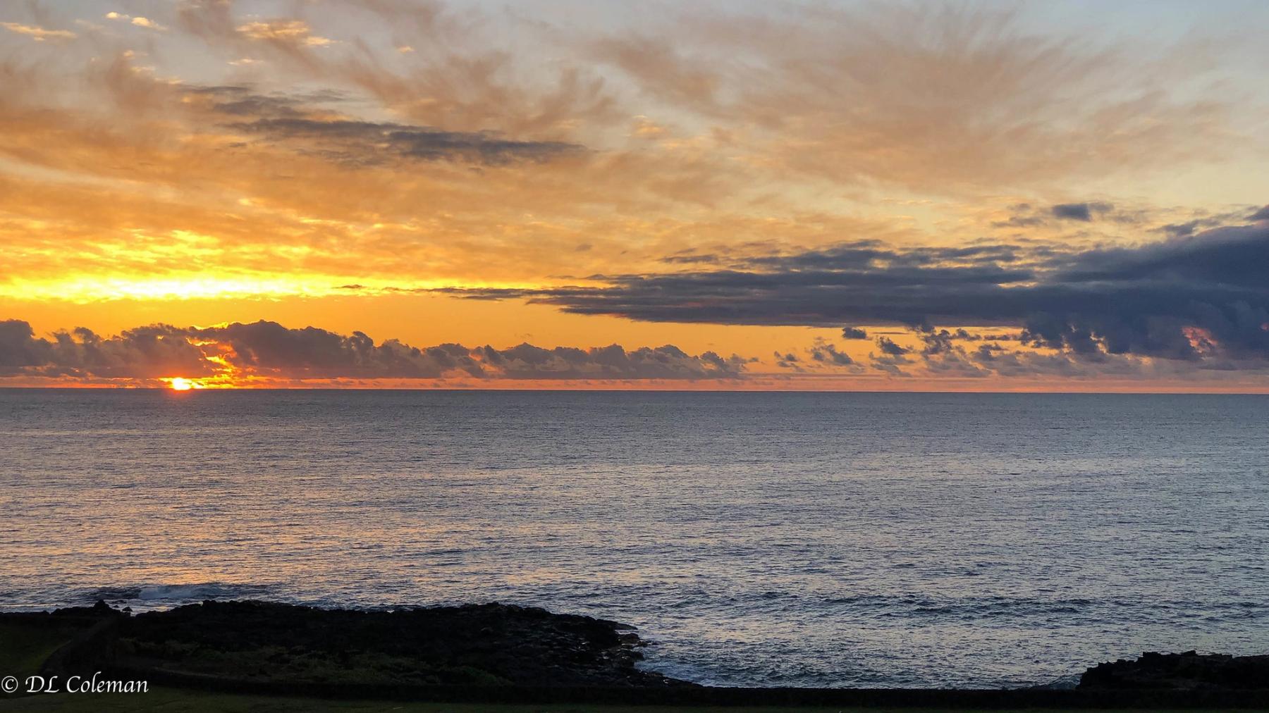 Golden sunrise breaking through layered clouds over a calm ocean horizon with dark rocky shoreline in the foreground