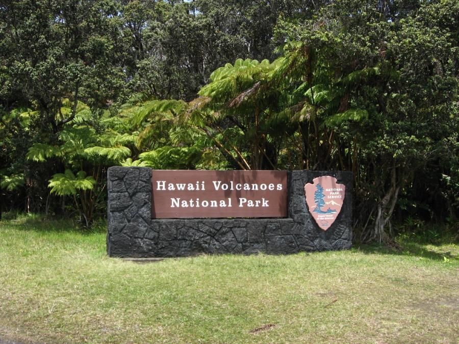 Entrance sign for Hawaiʻi Volcanoes National Park set in a stone wall with ferns and dense forest behind.