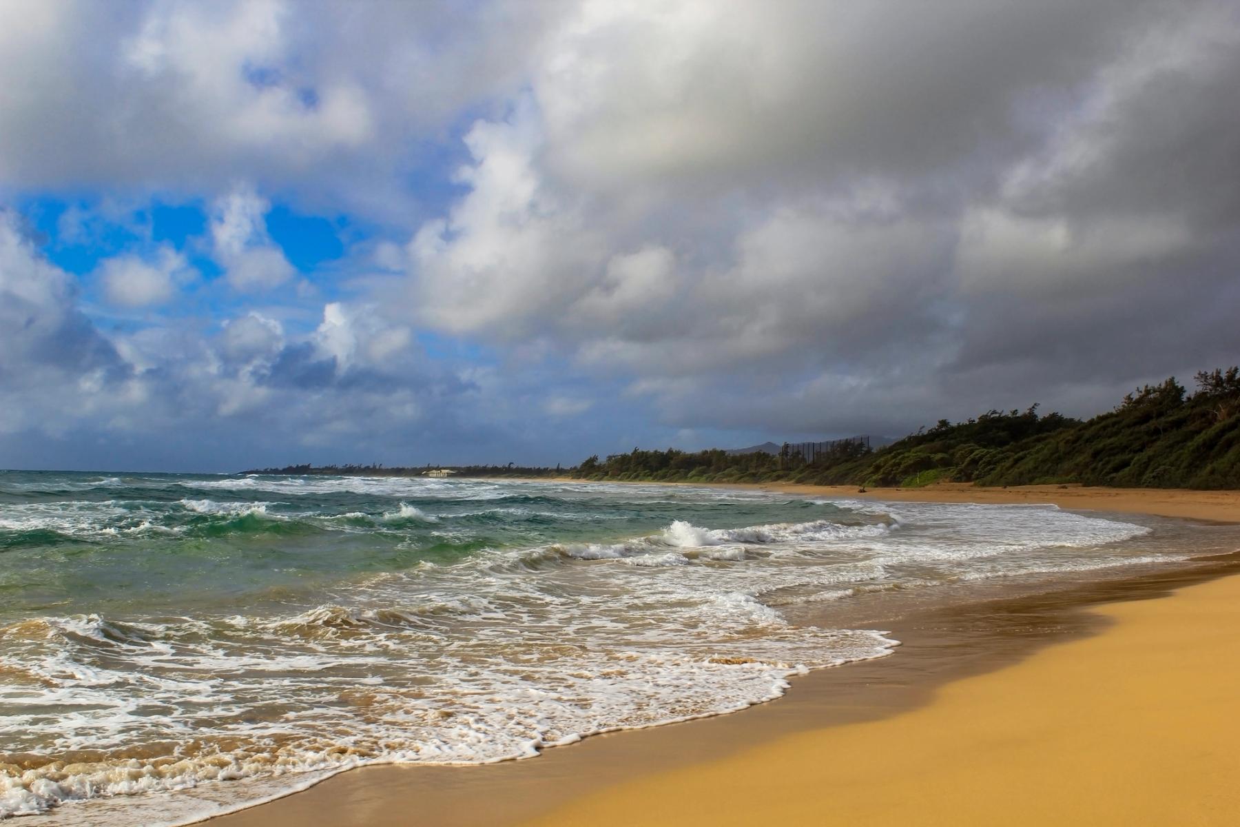 Curving sandy beach with foamy waves, green-blue water, and dramatic clouds along a tree-lined Kauaʻi coastline