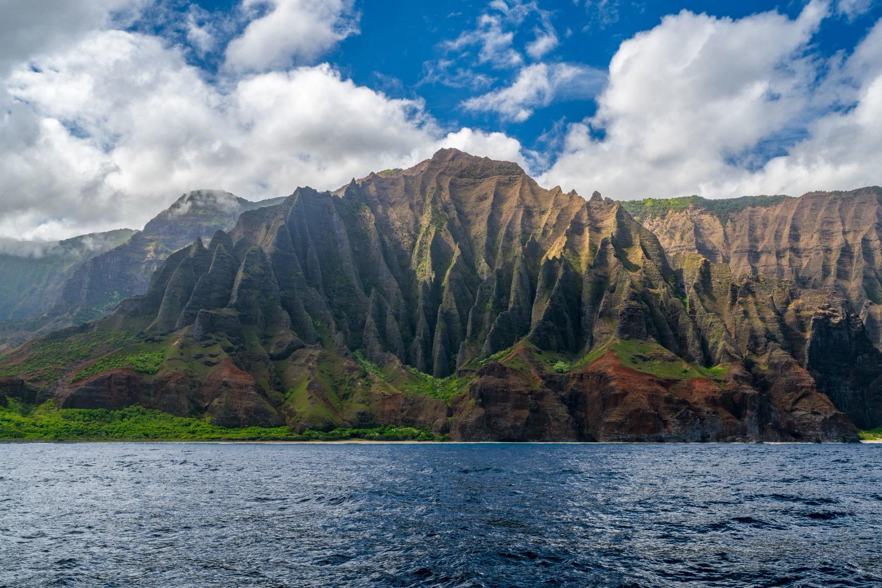 Towering ribbed Nā Pali sea cliffs on Kauaʻi rising above a choppy blue ocean under bright clouds