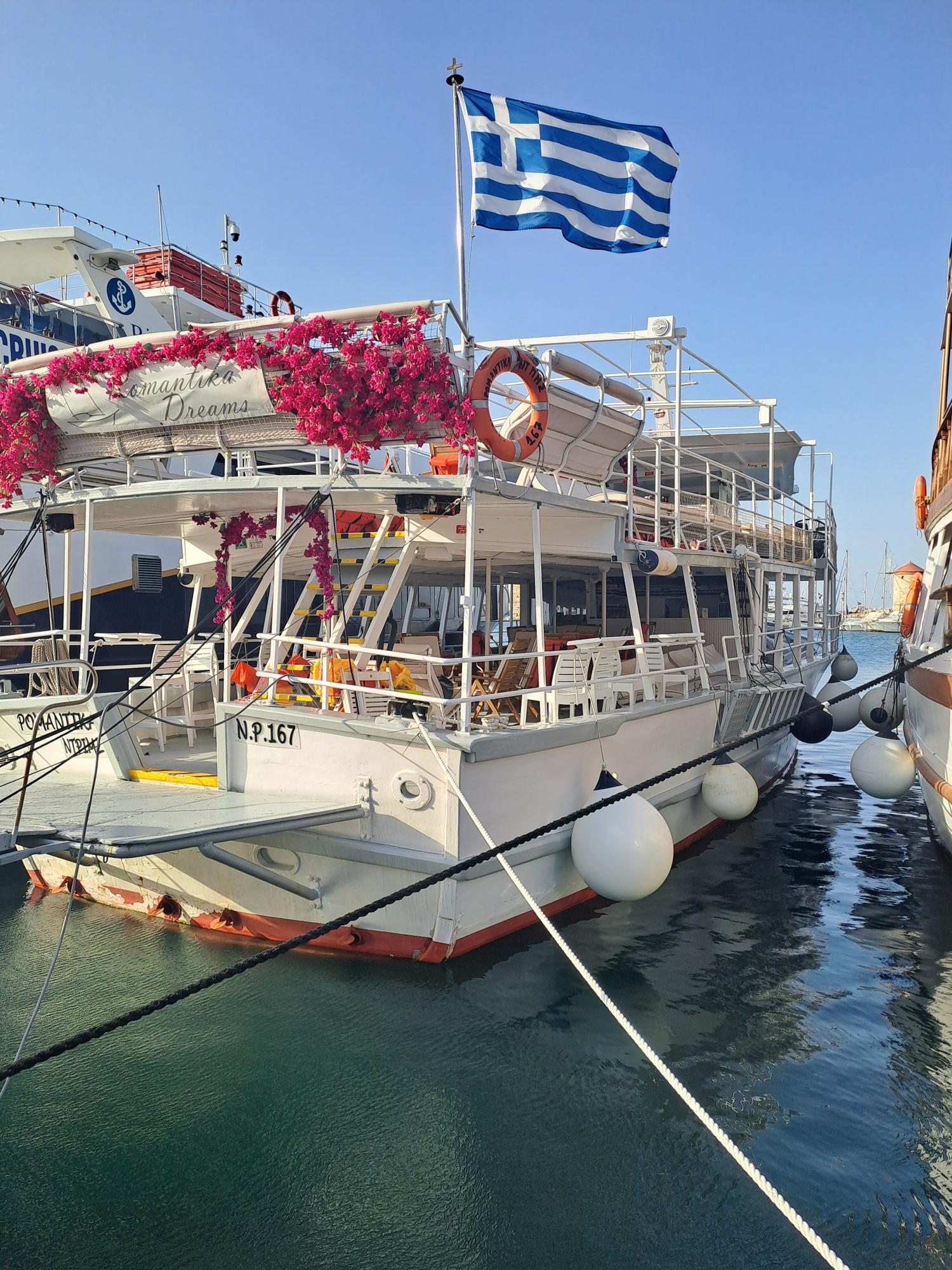White excursion boat decorated with pink flowers and flying a Greek flag at the harbour.