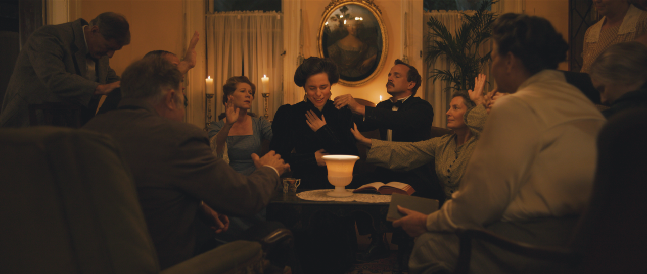 Men and women in a lamplit parlor lay hands on a woman in prayer