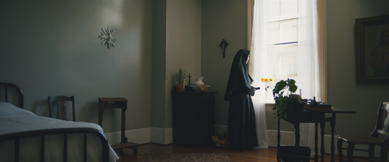 A religious sister stands at a sunlit window in a quiet bedroom