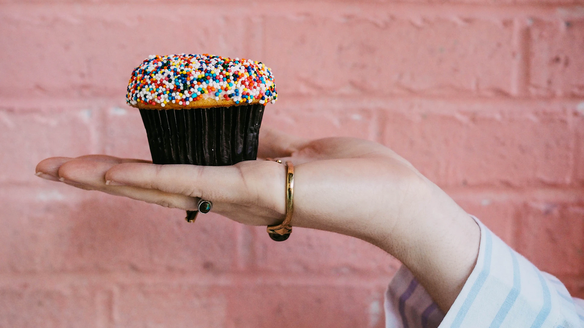 A hand holding out a small cupcake covered in a colourful hundreds and thousands topping