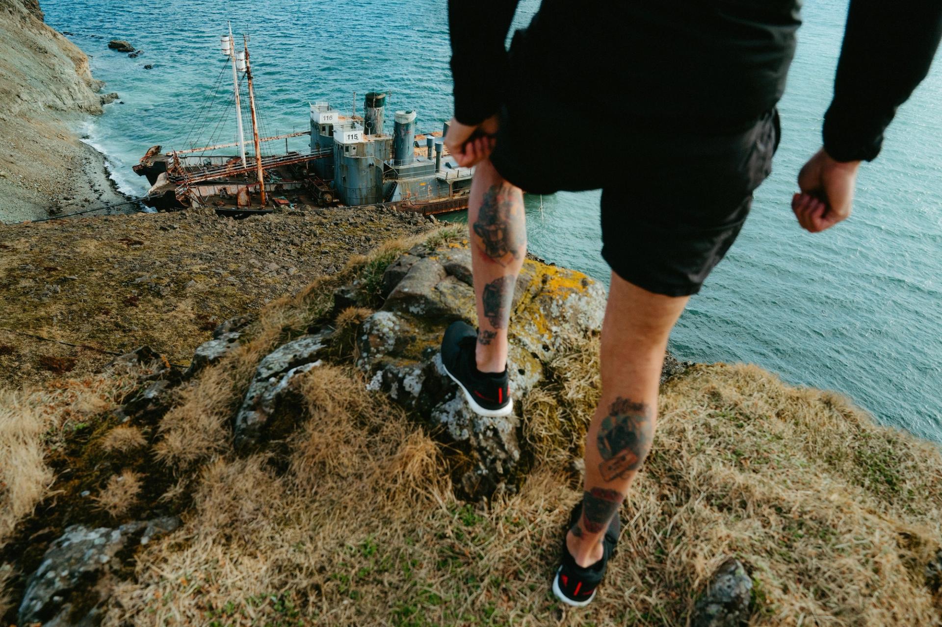 Runner's tattooed legs above a rusted shipwreck on the coast — Björn Lexius, Iceland