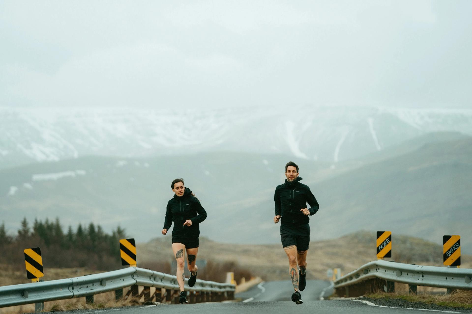 Two runners on a mountain road with snow-capped peaks behind — Björn Lexius, Iceland