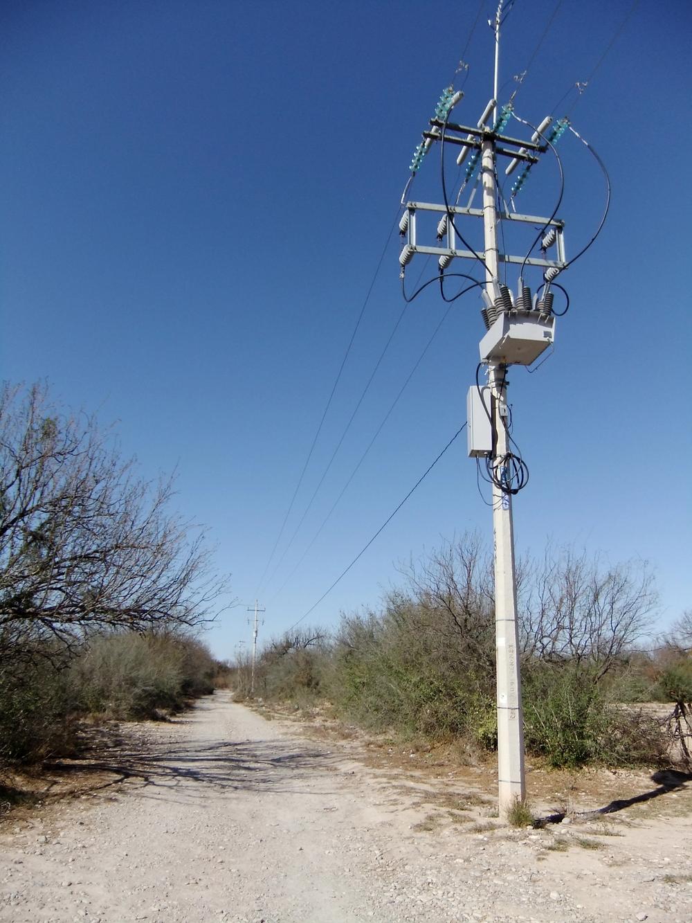 Pole mounted NOJA Power OSM Recloser Installation on the side of gravel road, with clear blue skies and shrubbery lining the road