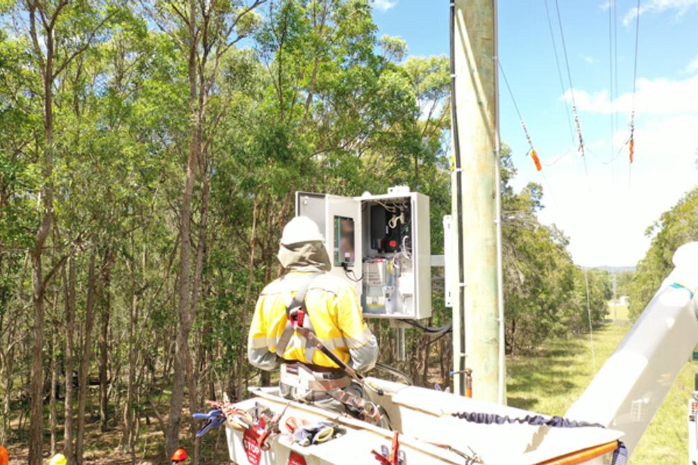 A line worker is in a crane facing the NOJA Power RC20 control cubicle under installation in Queensland Australia. The background has trees and light blue skies.