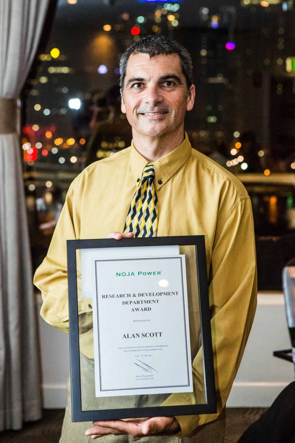 Man wearing a yellow button up shirt and black and yellow tie, holding a certificate with city lights behind