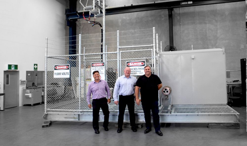 Left to Right, Dean van Wijk, Neil O’Sullivan and Julian Rauwendaal are standing in front of a testing cage in a warehouse with a NOJA Power GMK mobile substation unit behind