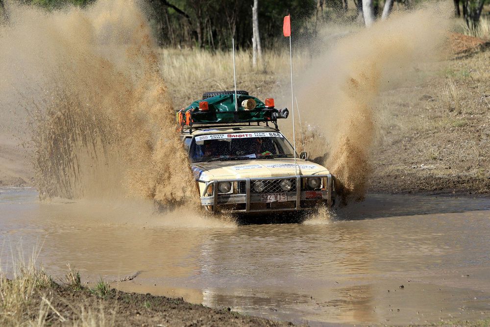 4WD rally car driving through mud with water spraying up both sides of the car 