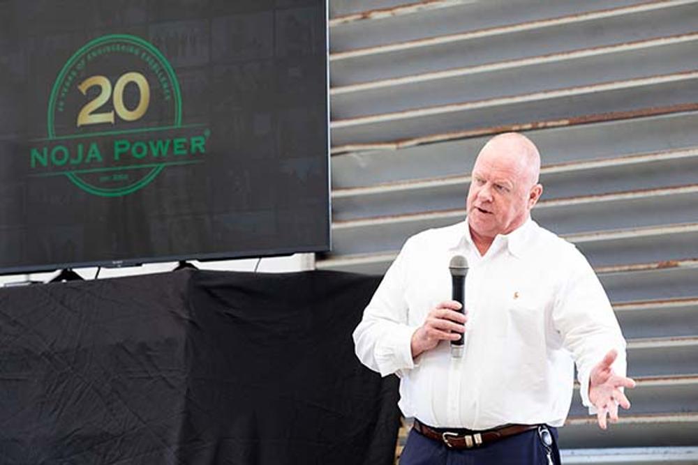 Man wearing white shirt speaking into a microphone, with TV to the left displaying logo with 20 in old and NOJA power in green below 
