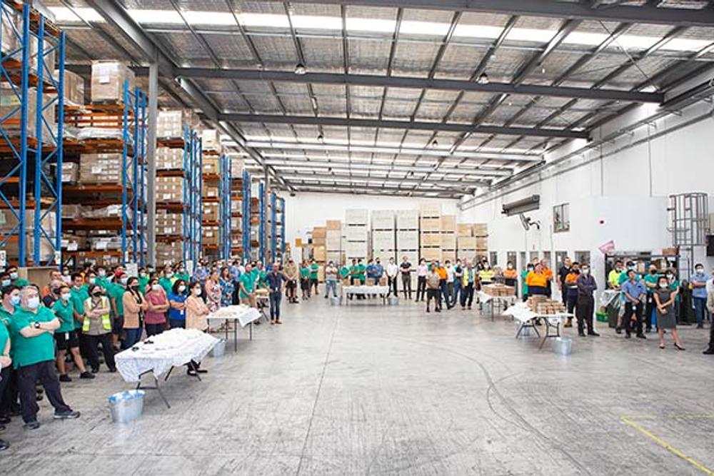 Large group of people standing around the edges of warehouse behind trestle tables