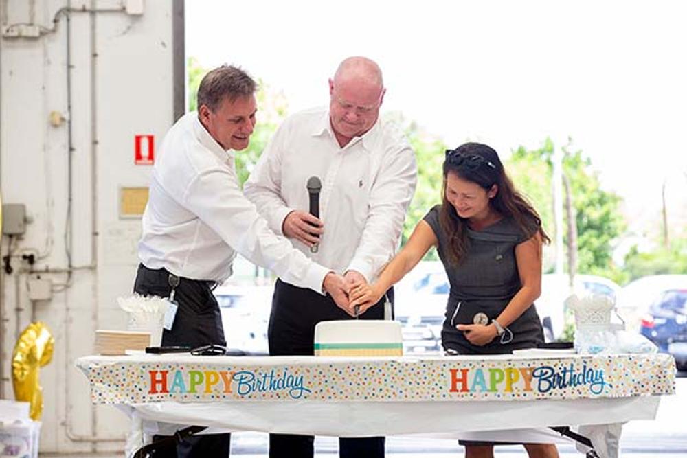 Three people, two men and one female are holding a knife together cutting a white cake on table with 'Happy birthday' banners below 