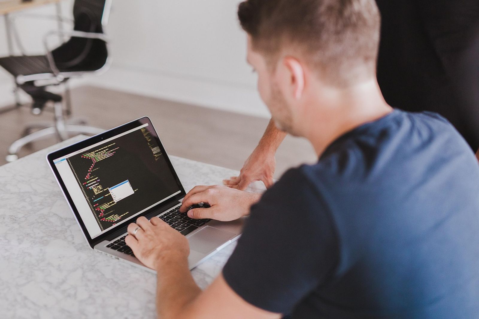 A man in a dark blue t-shirt types on a laptop showing code while another person leans on the marble desk beside him
