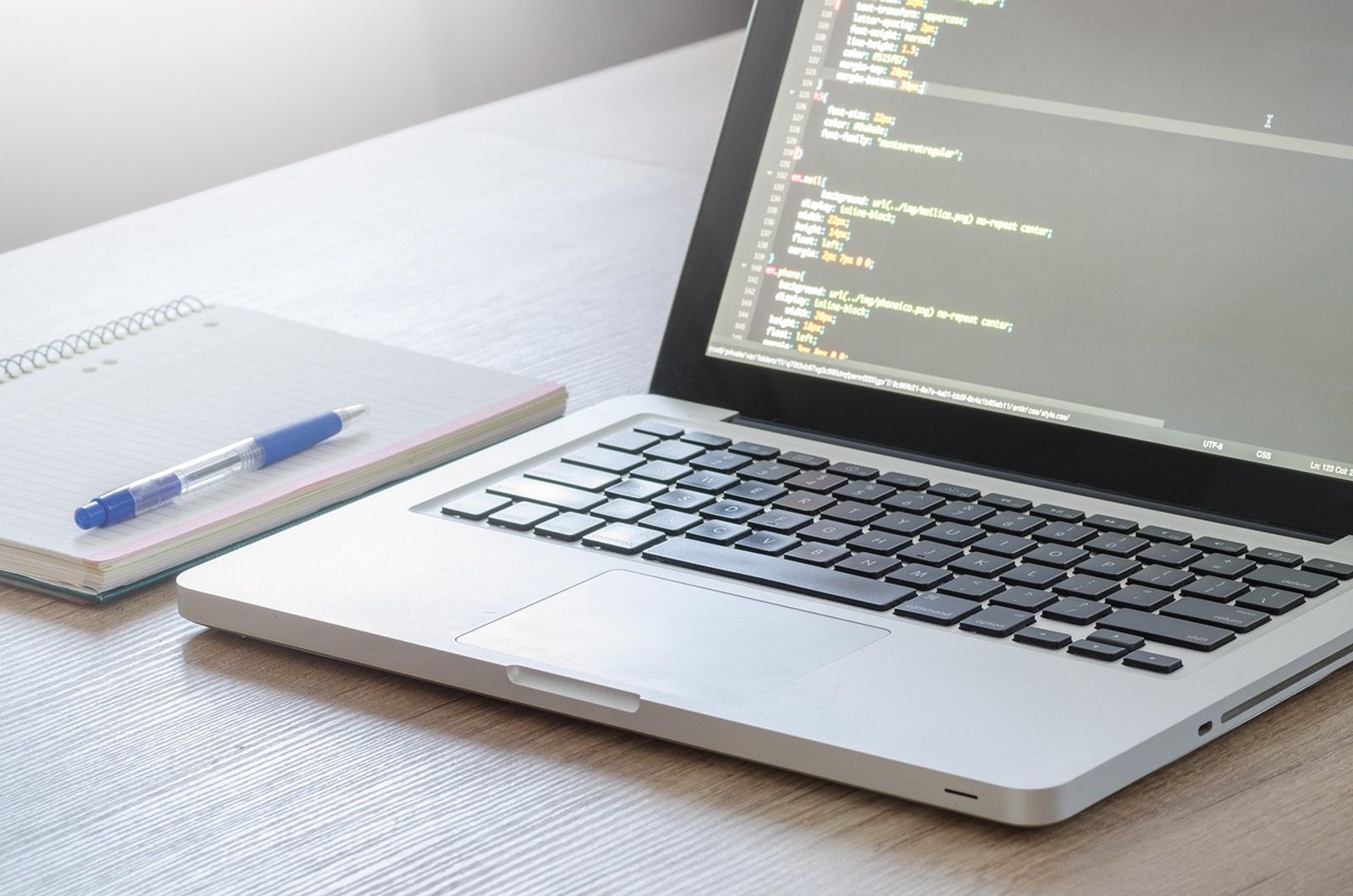 Silver laptop showing lines of CSS code on screen beside a spiral notebook and blue pen on a wooden desk