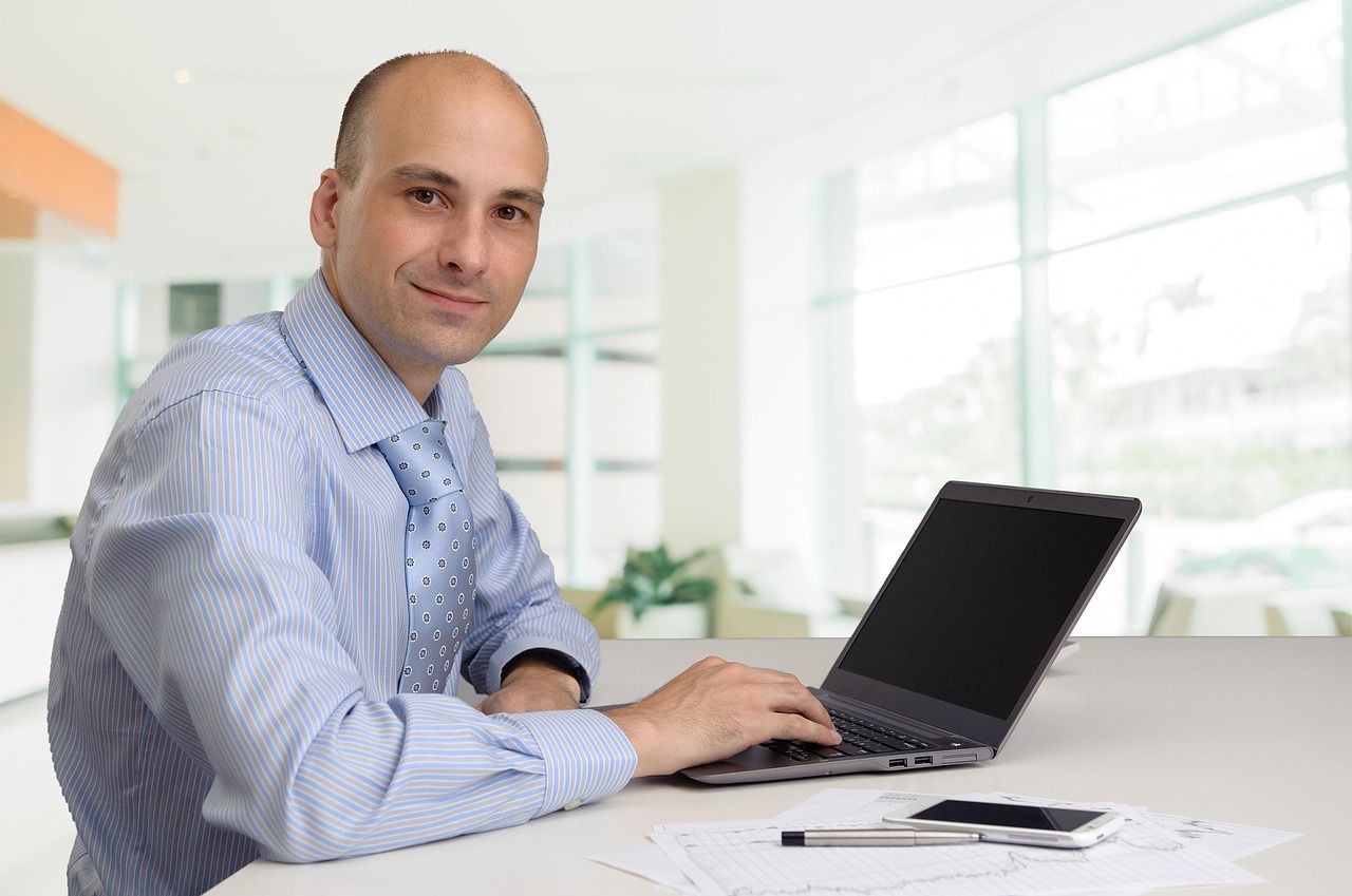 Smiling man in a blue striped shirt and tie sits at a white desk with a laptop, papers, a phone and a pen
