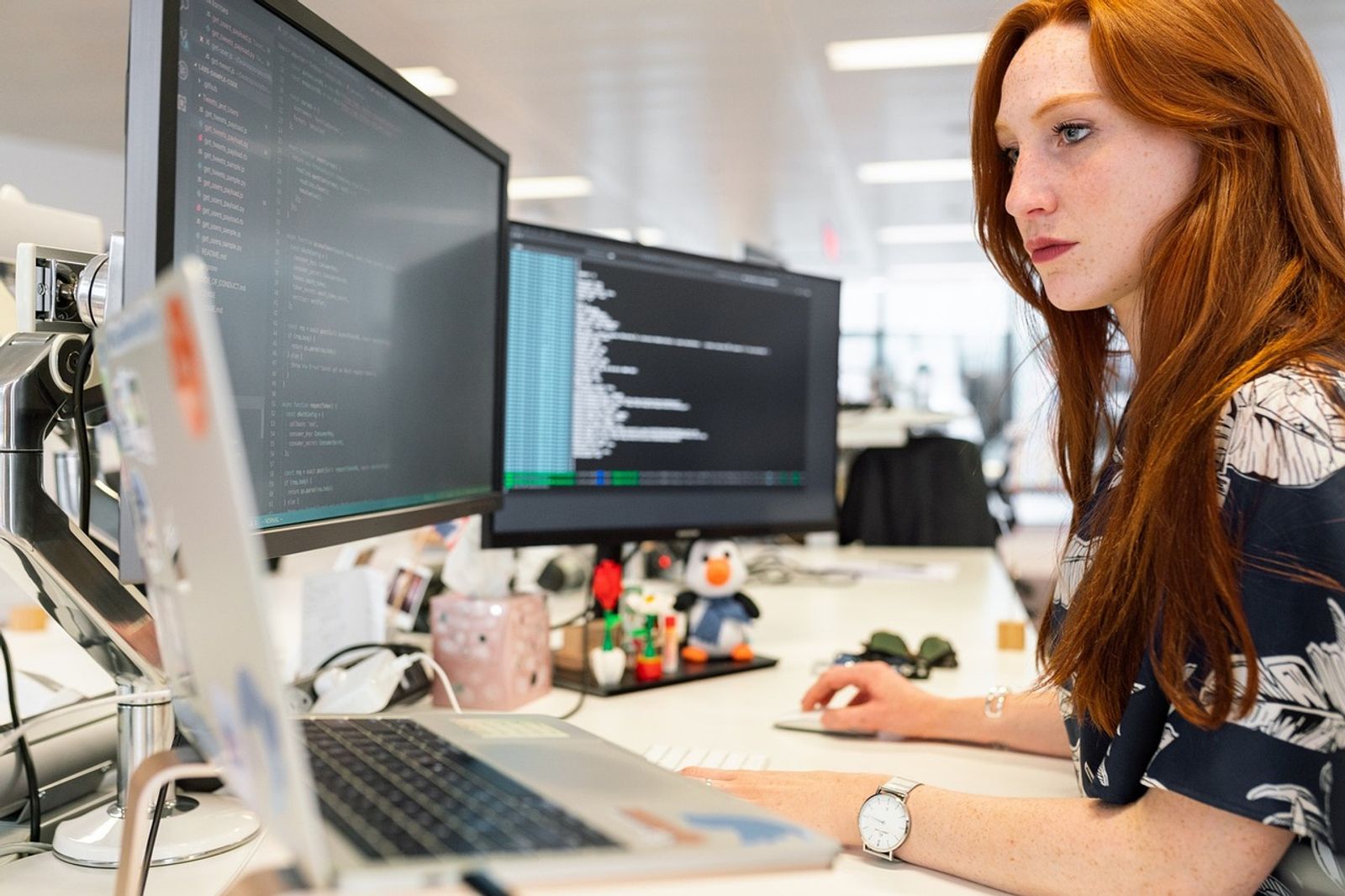 Woman with long red hair working at a desk with two monitors showing code and a laptop in an open office