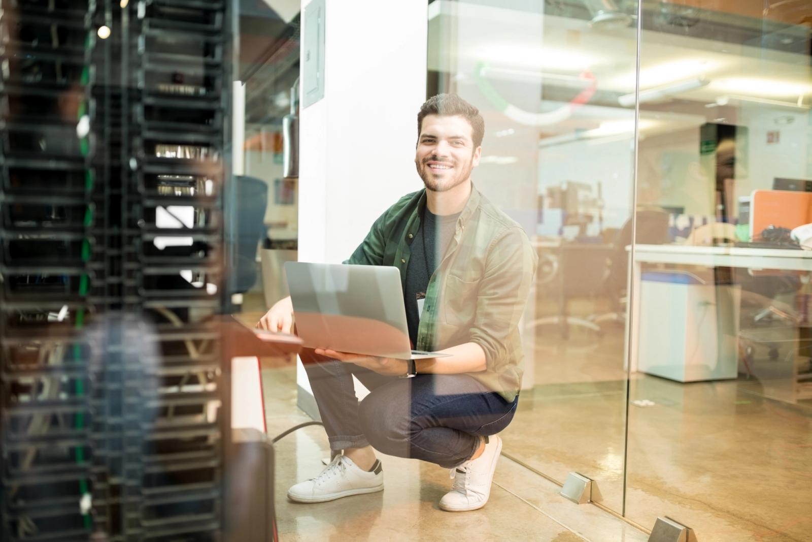 SAP integration engineer holding a laptop beside server racks in a data center with an office behind glass