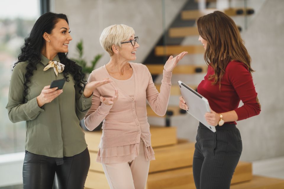 Three women stand talking in a bright modern office; one holds a phone, another a tablet, near a wooden staircase