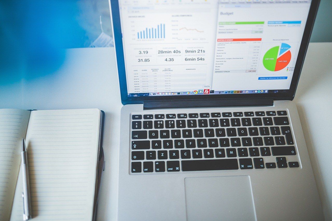 Laptop on a white desk showing analytics charts and a budget pie chart, beside an open lined notebook and pen