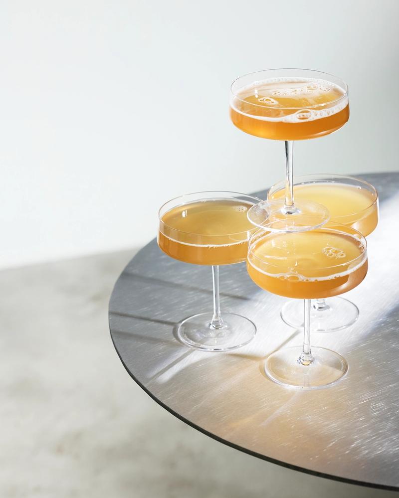 Four coupe glasses filled with golden, alcohol-free Villbrygg served on a brushed metal table in a minimalist setting.