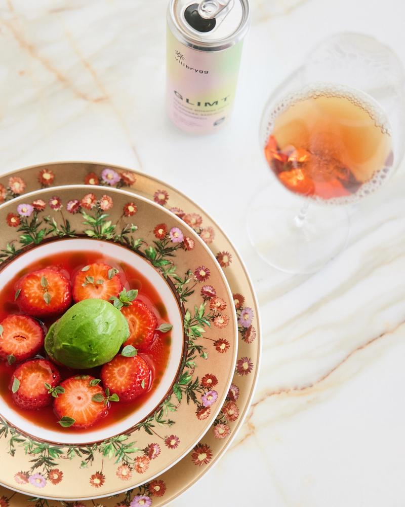 Strawberries and green herb sorbet served in a floral, gold-rimmed bowl, with Villbrygg Glimt in a glass and can on a marble table.