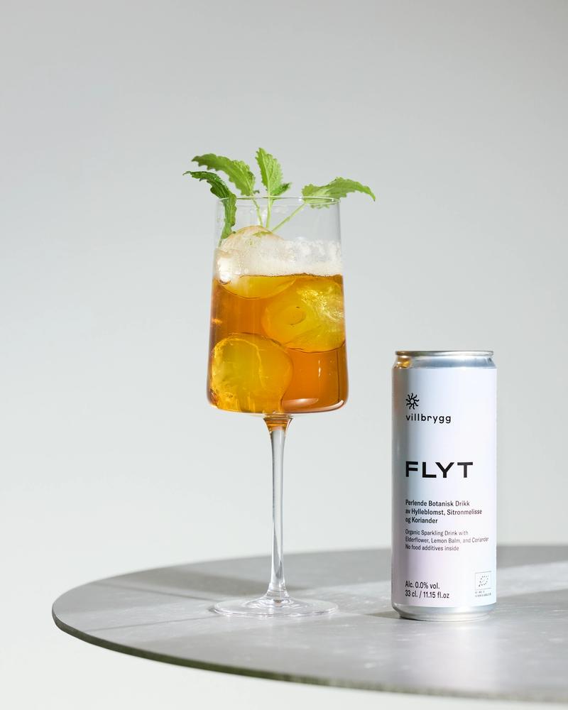 Villbrygg FLYT served over ice with fresh herbs in a wine glass, next to a silver can, placed on a round table.