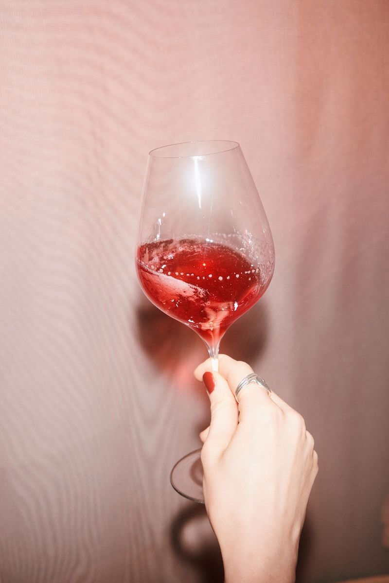 A hand holding a wine glass with Villbrygg Fjell – a deep red, non-alcoholic drink – against a soft pink background.
