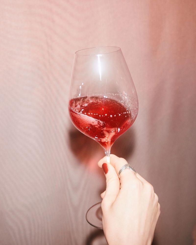 A hand holding a wine glass with Villbrygg Fjell – a deep red, non-alcoholic drink – against a soft pink background.