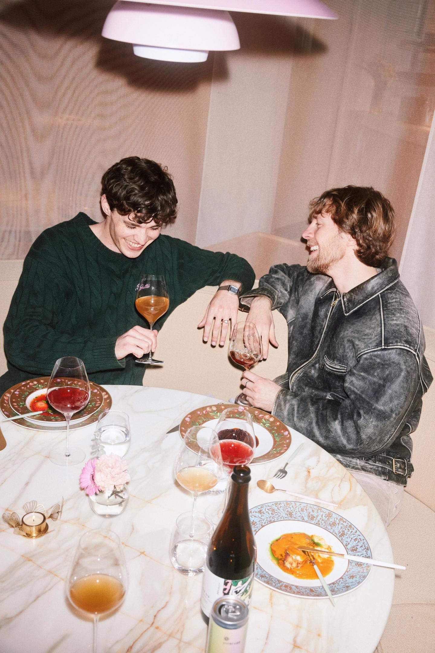 To venner ler sammen mens de nyter alkoholfrie drikker fra Villbrygg rundt et rundt marmorbord, dekket med blomstrete tallerkener og retter fra restauranten Mon Oncle. En flaske Villbrygg står midt på bordet, omgitt av vinglass og lun stearinbelysning.