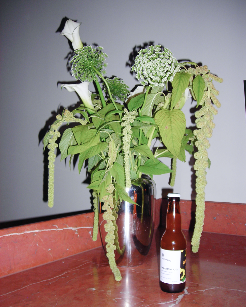 Villbrygg Hagen placed next to a green, sculptural floral arrangement in a silver vase on a red marble ledge.