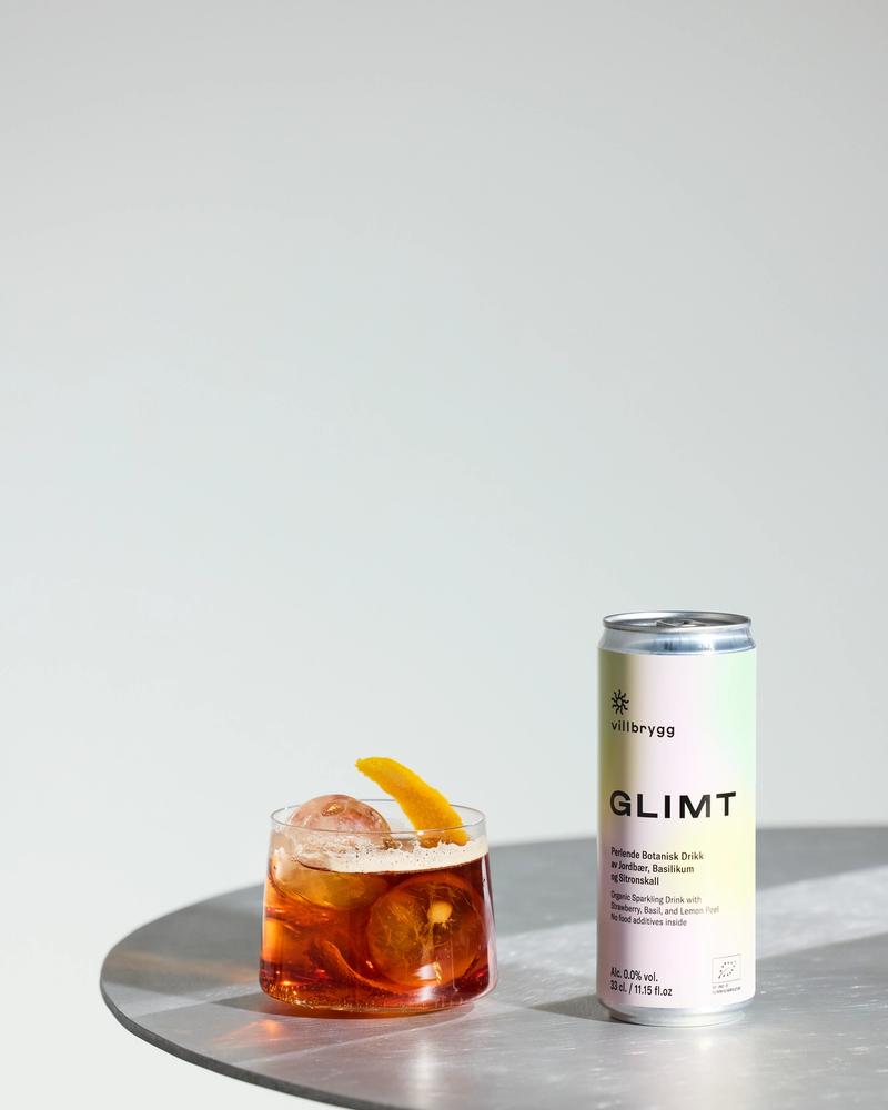 Villbrygg GLIMT served in a low glass with ice and citrus peel, next to a pastel-colored can on a round metal table.