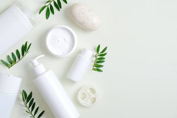 White body wash bottles and cream jar with green leaves on a pale background