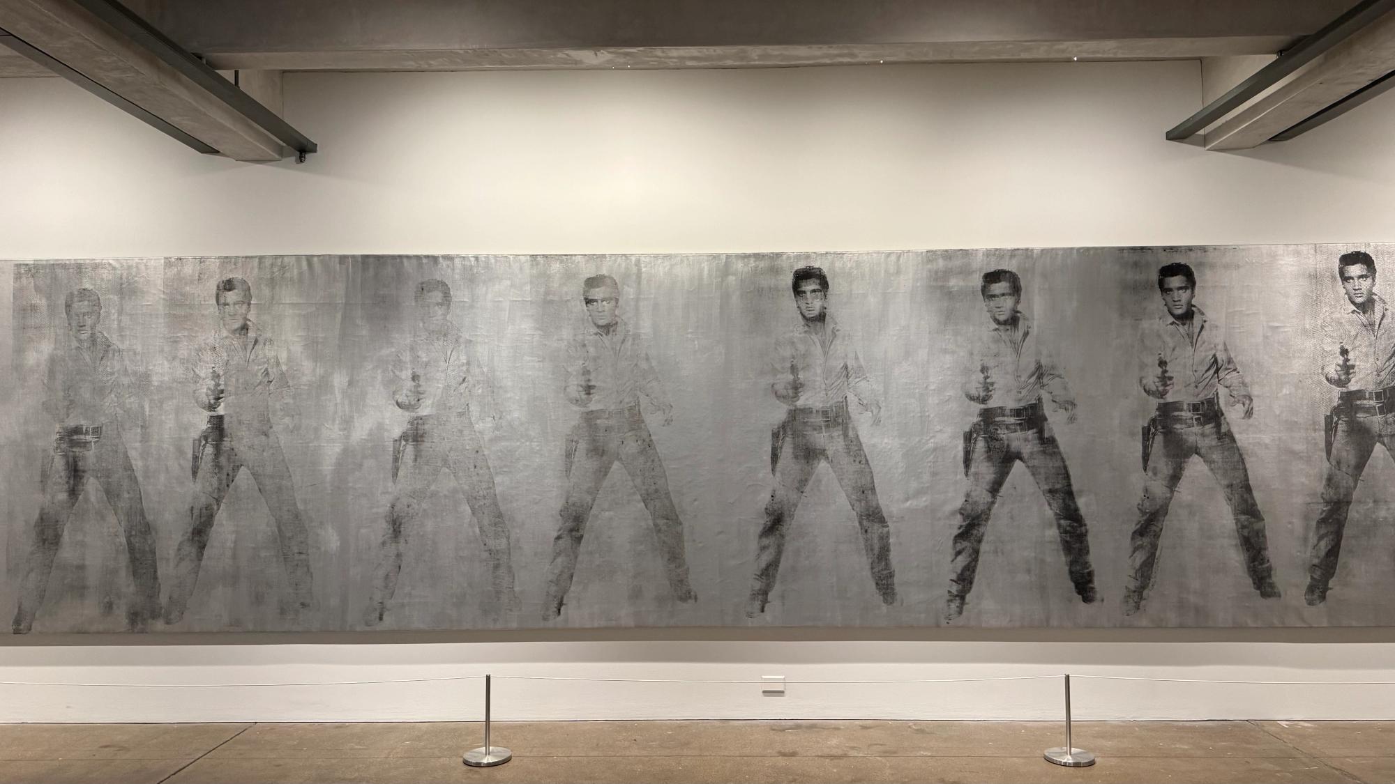 Large silk-screen painting with many images of Elvis Presley in a gunslinger pose, on a shiny silver background