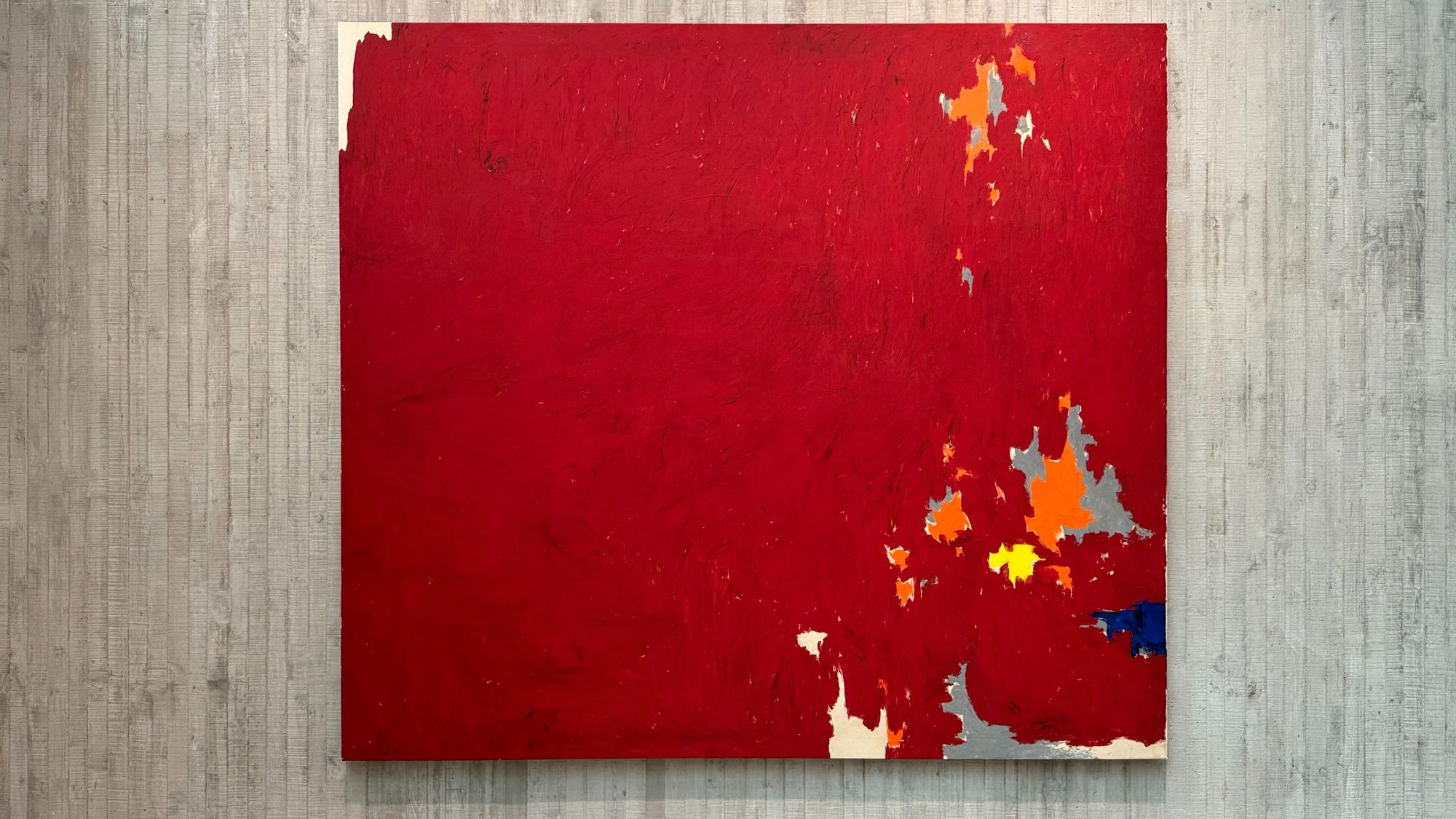 A mostly-red paitning with patches of grey and orange. By Clyfford Still.