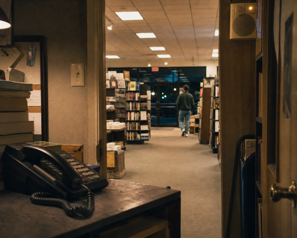 A black corded office phone beside stacks of books, looking out from a bookstore office toward an anonymous person walking to the exit at night.