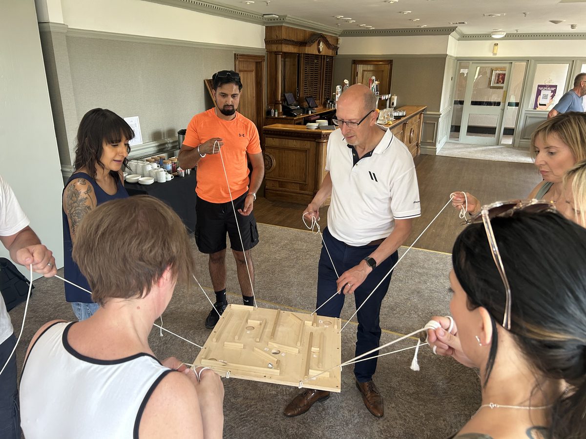 A team of six guiding a ball through a wooden maze on strings