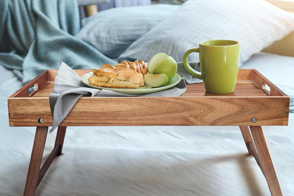 A wooden serving tray on white bedsheets holding fresh croissants, fruit, and a hot cup of coffee.