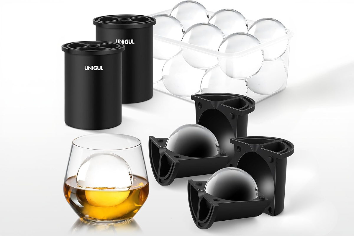 A large, crystal-clear spherical ice ball melting slowly inside a premium crystal lowball glass filled with an amber craft cocktail on a dark marble surface.