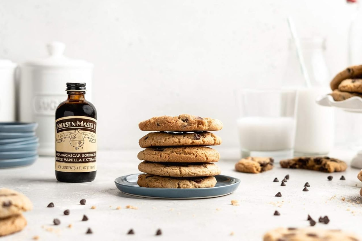 An elegant dark amber glass bottle of Nielsen-Massey Madagascar Bourbon Pure Vanilla Extract sitting on a modern marble countertop next to a stack of freshly baked chocolate chip cookies