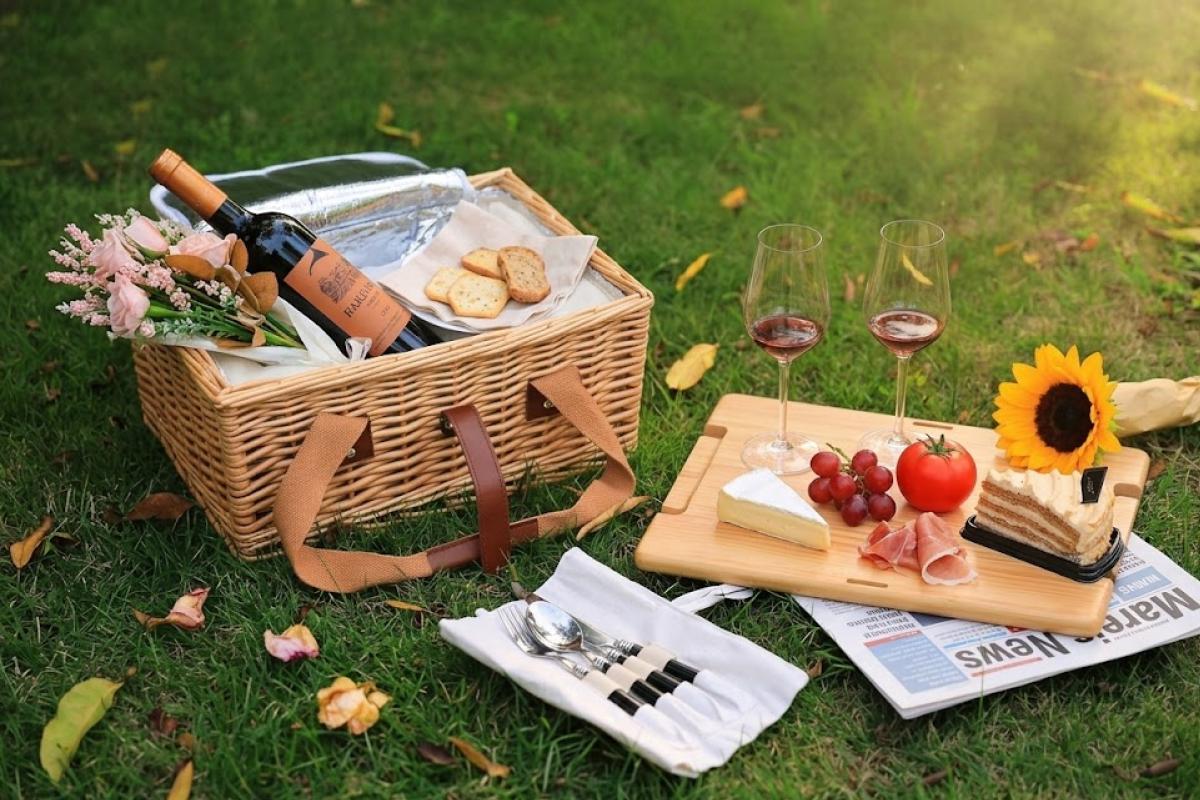 A handwoven picnic basket in wicker next to a board of charkuterie and two glasses of wine.