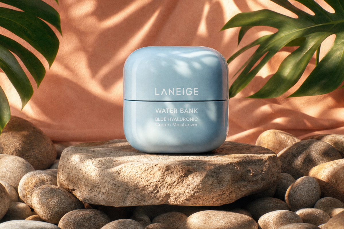A professional product photograph of a pastel blue, rounded square jar of Laneige Water Bank Blue Hyaluronic Cream Moisturizer. The jar sits prominently on top of a cluster of smooth, rounded river stones. In the background, large, vibrant green tropical split-leaf philodendron leaves fan out against a warm peach-toned backdrop. Soft, artistic shadows of palm fronds drape across the scene, creating a sun-drenched, tranquil atmosphere.