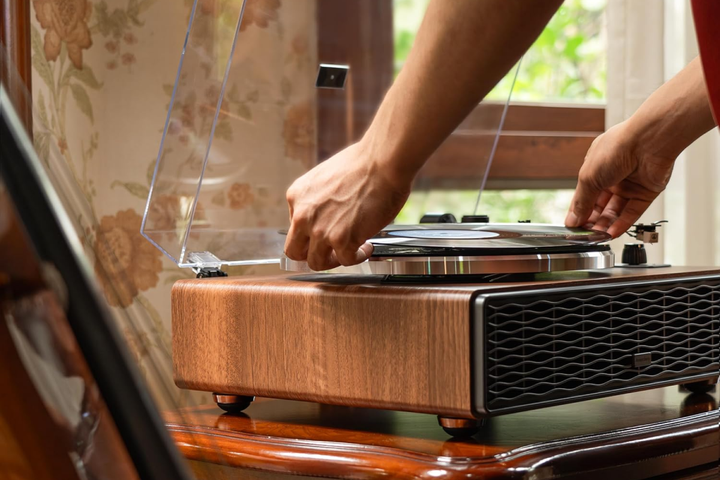 Un tocadiscos de estilo vintage con base de madera girando un disco de vinilo junto a una pequeña pila de portadas de álbumes clásicos en un estante de mediados de siglo.