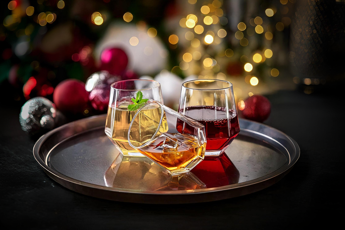 A pair of elegant, crystal-clear stemless wine glasses filled with a rich red wine, resting on a polished dark wood table bathed in soft, christmas lights