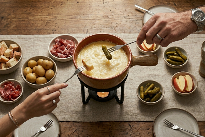 Un pot à fondue lumineux entouré de petits bols contenant des pommes de terre, des cornichons, des pommes et du pain artisanal en cubes.