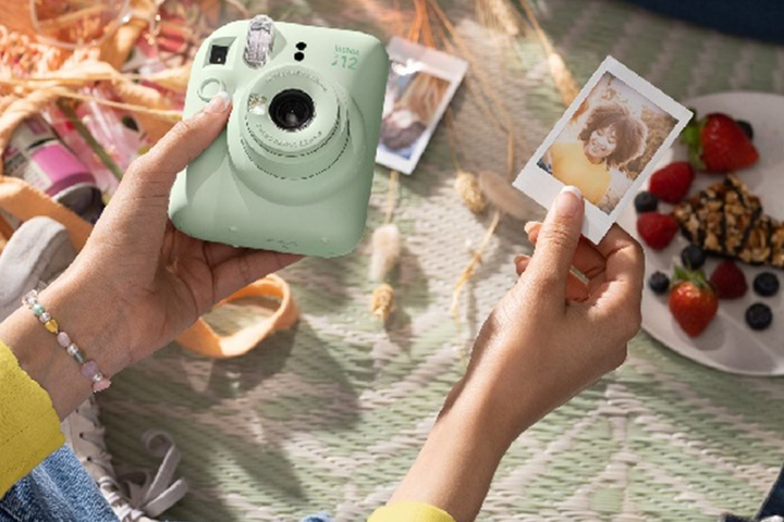 A pastel mint green Fujifilm Instax Mini 12 instant camera sitting next to a scattered pile of credit-card-sized color photos.