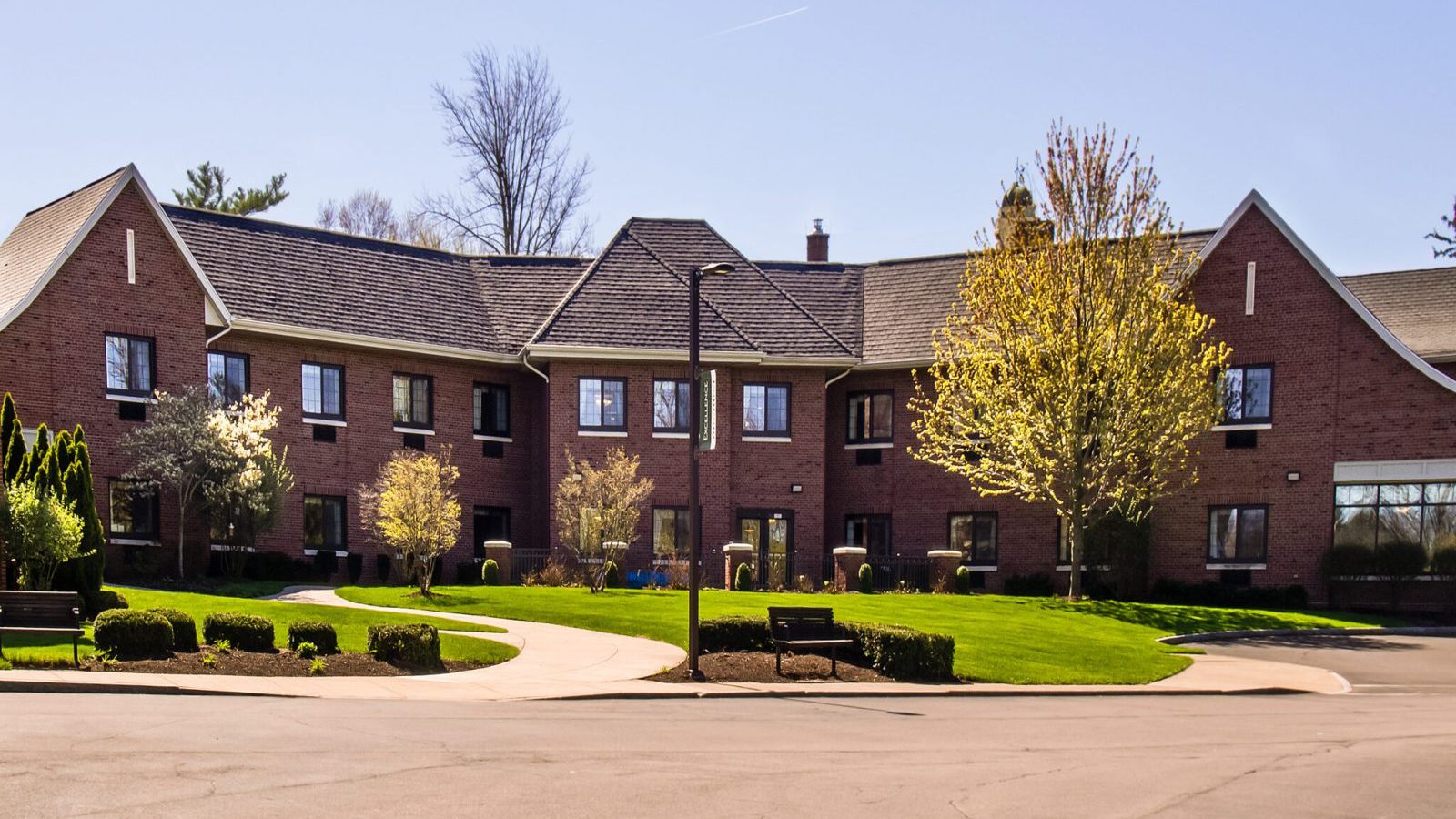 Exterior view of a modern assisted living community with a well-maintained brick building, landscaped gardens, walking paths, and a welcoming entrance, providing a safe and comfortable environment for seniors.