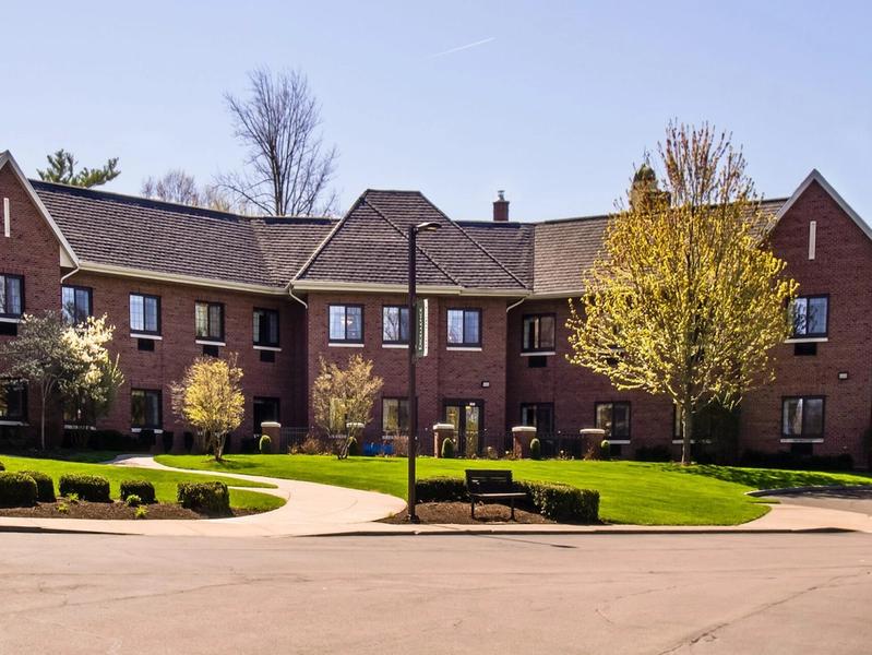 Exterior view of a modern assisted living community with a well-maintained brick building, landscaped gardens, walking paths, and a welcoming entrance, providing a safe and comfortable environment for seniors.